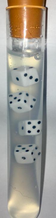 Four white 1mm plastic dice suspended at different heights inside a sealed test tube of clear Ibumix suspension, unmoved after more than two years